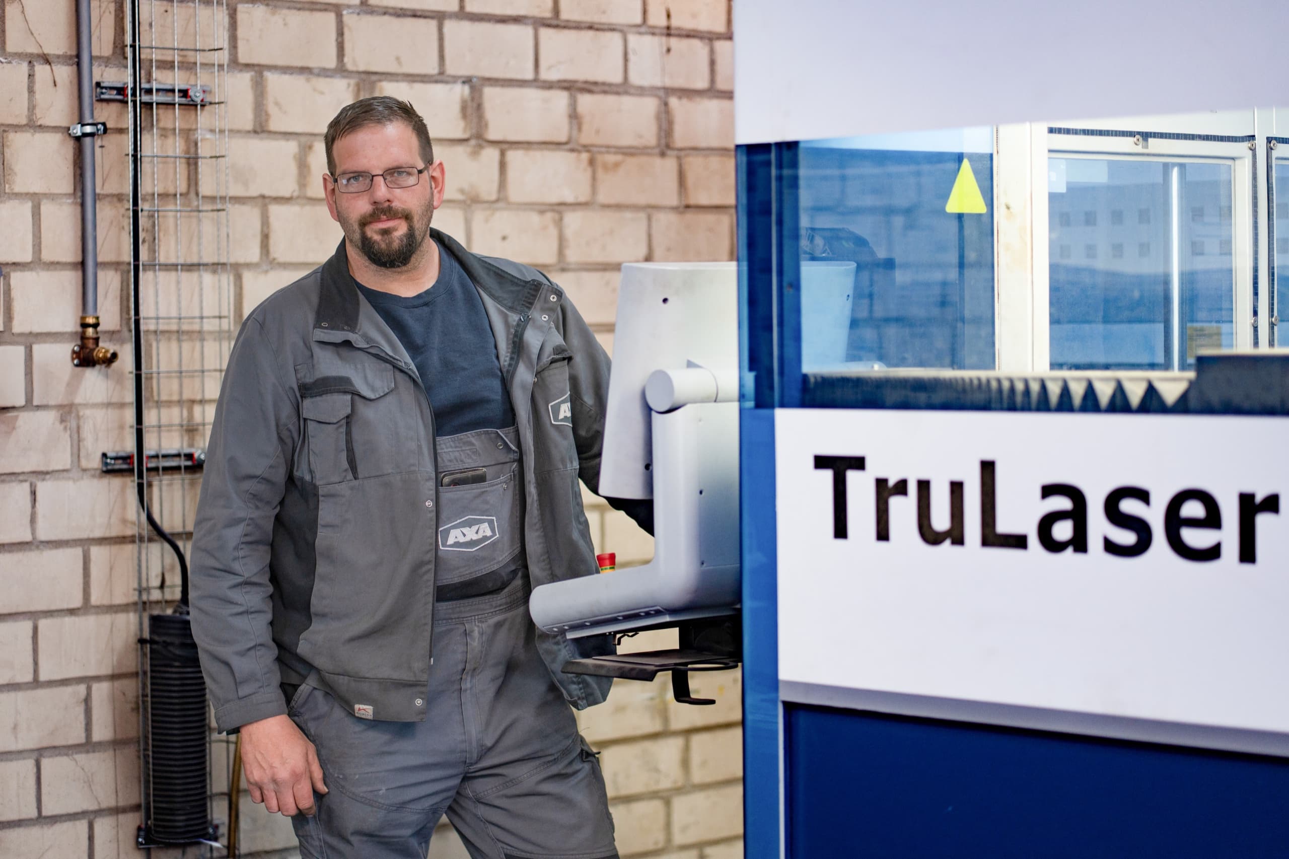 AXA Mitarbeiter in grauem Firmen-Arbeitsanzug vor TruLaser Maschine