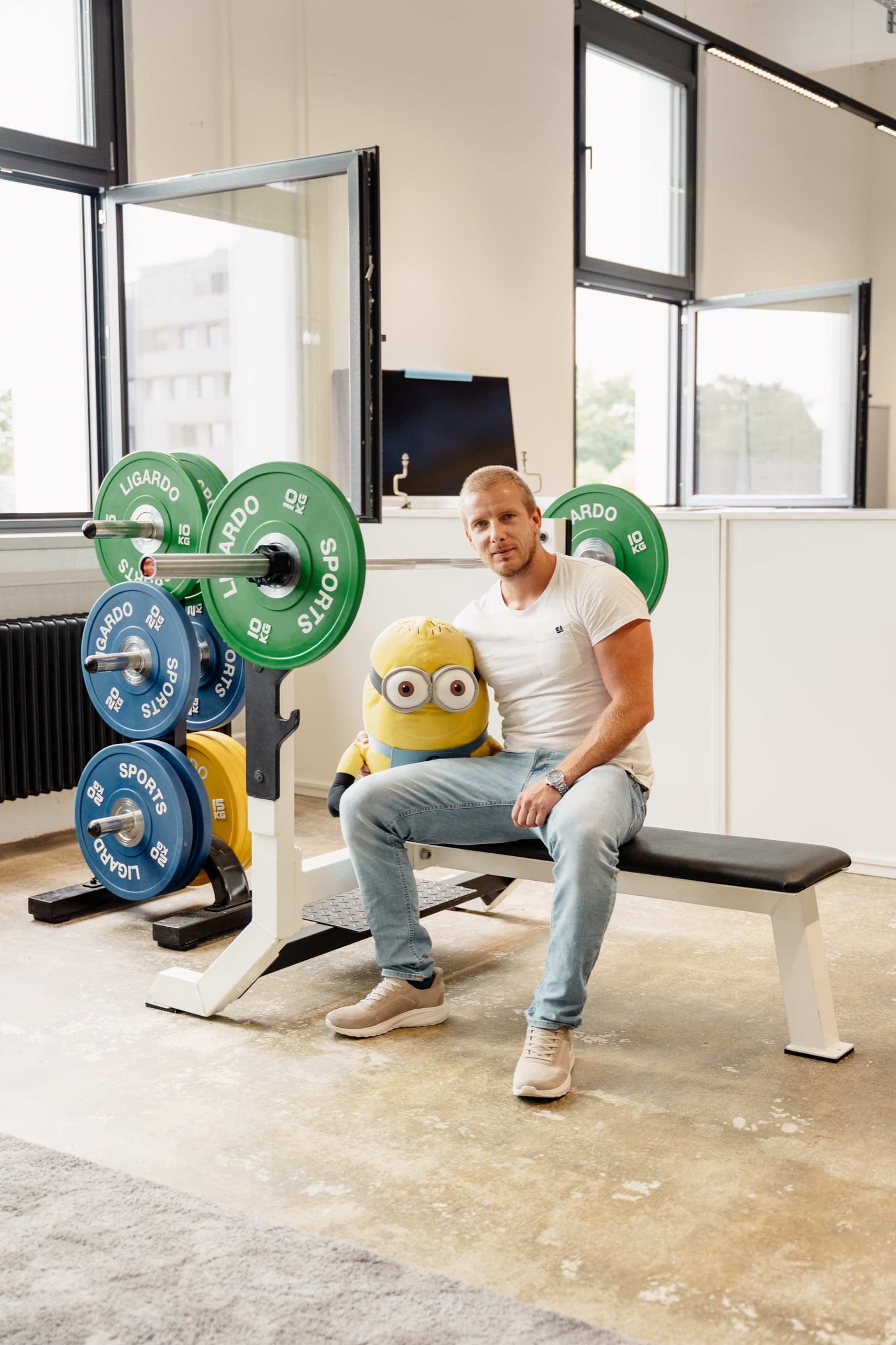 bee-doo Mitarbeiter sitzt im bürointernen Fitnessbereich mit Hantelständer und Minion-Maskottchen