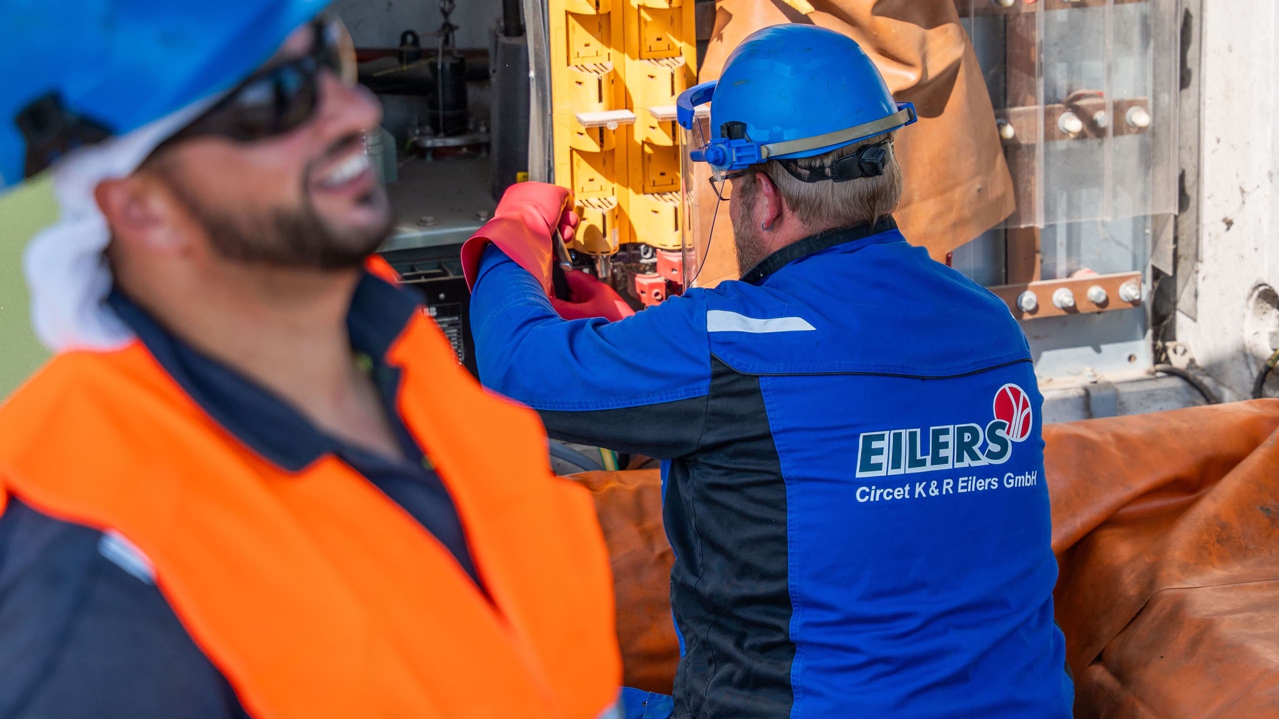 Elektriker mit EILERS-Jacke arbeitet an gelbem Kabelverteilerschrank, Kollege im Vordergrund