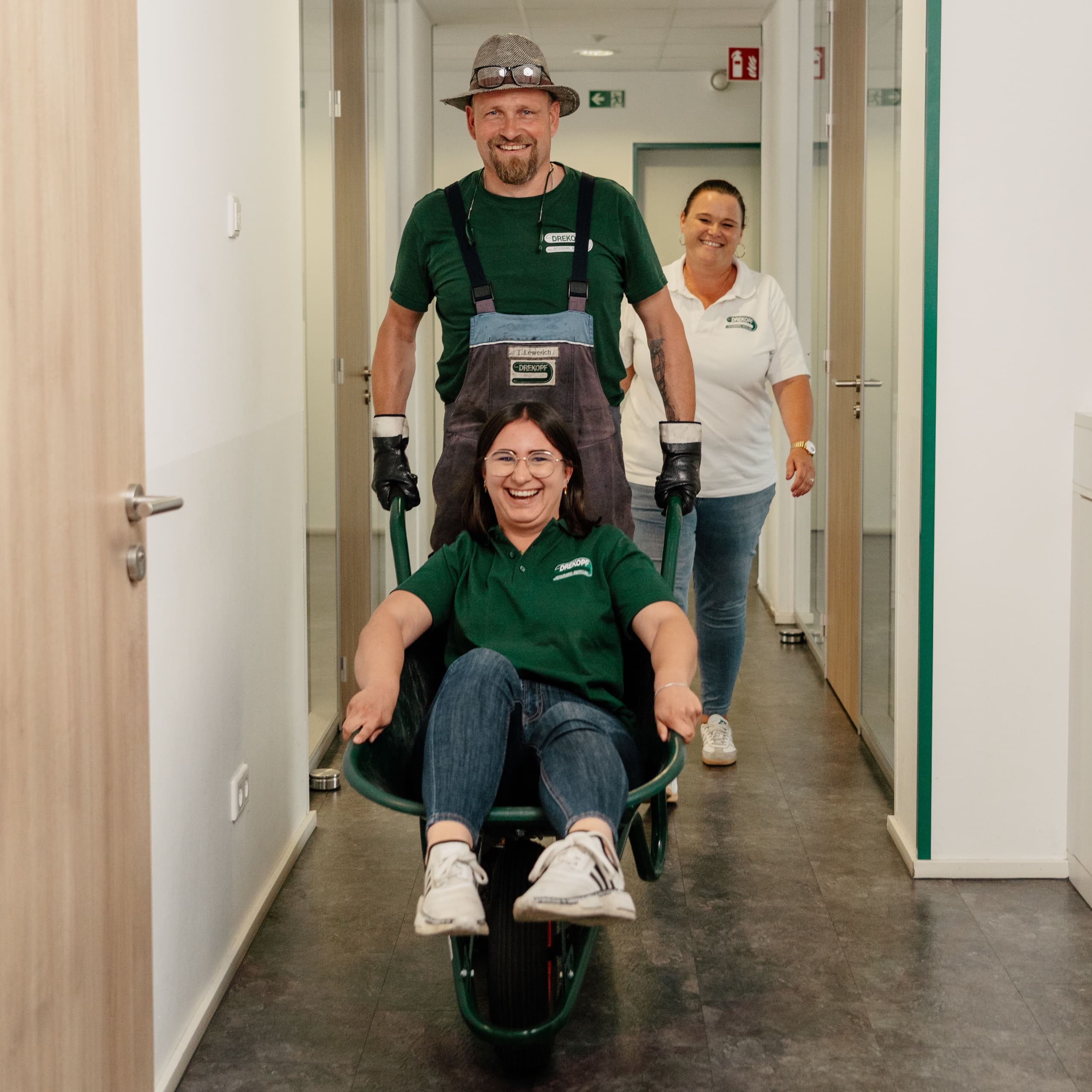 Drei Drekopf Mitarbeiter: Kollege schiebt lachende Kollegin in Schubkarre durch Büroflur – humorvolles Team-Foto