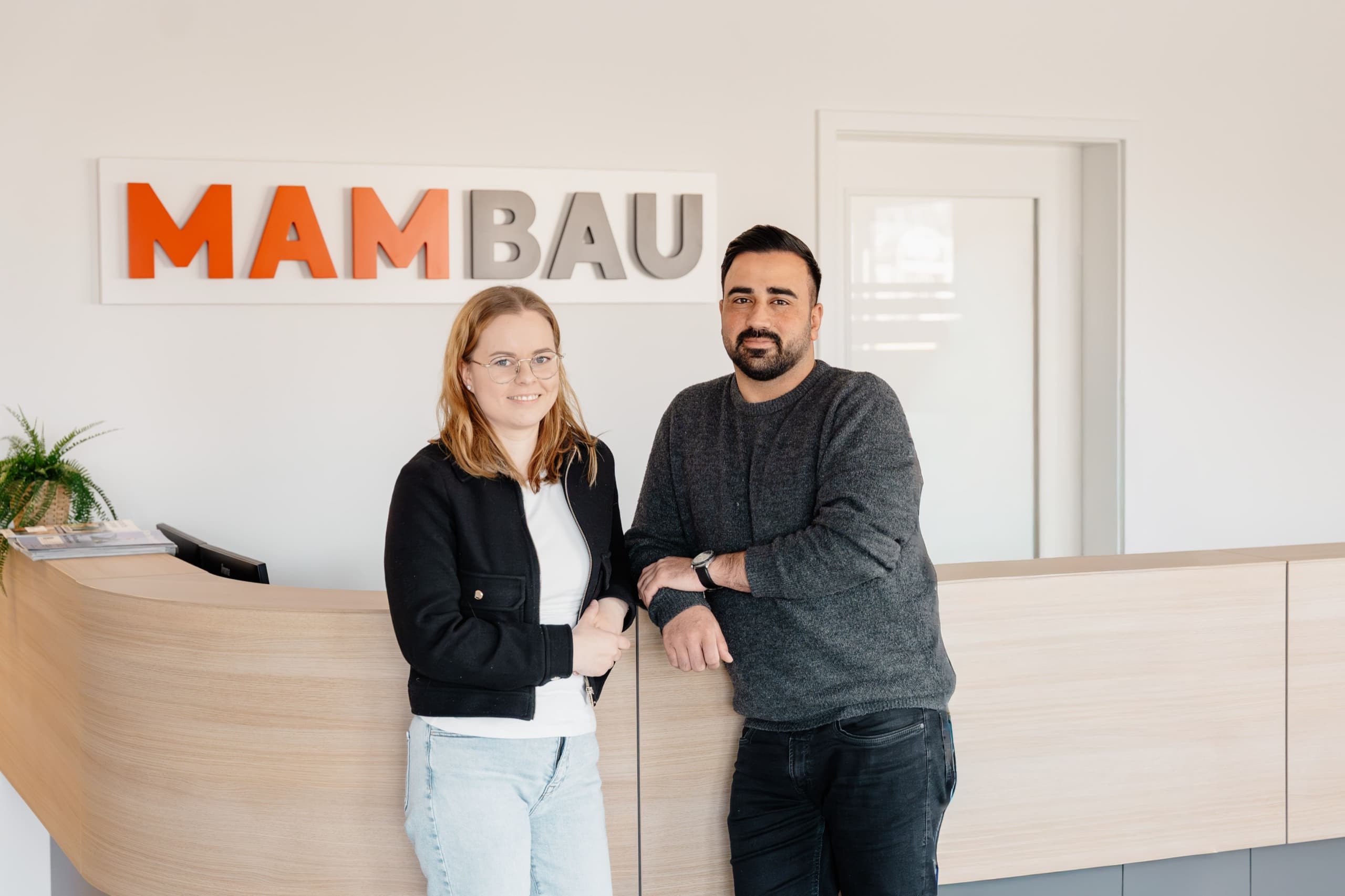 Zwei MAM-BAU Mitarbeitende an der Empfangstheke unter MAM-BAU Wandschriftzug