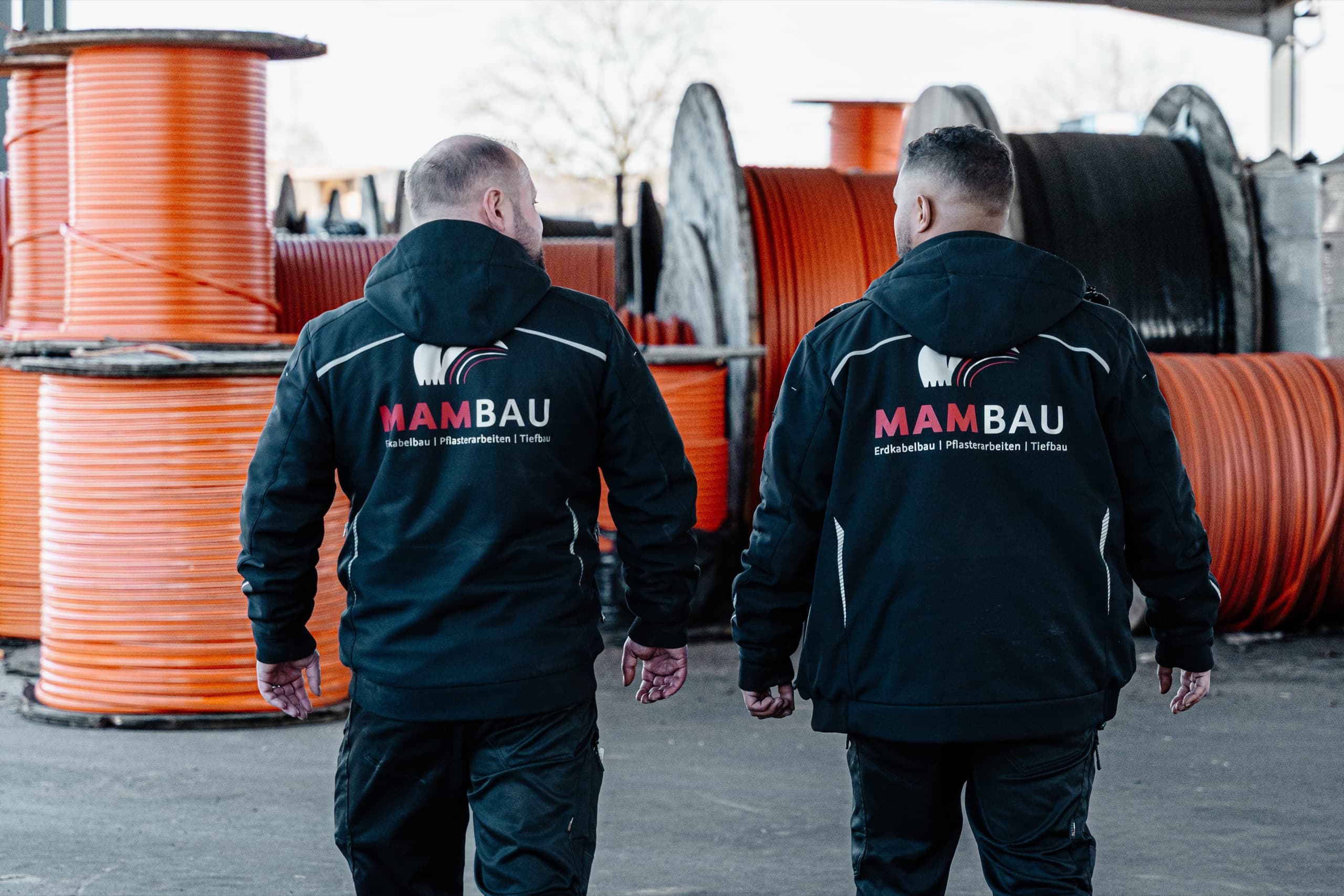Zwei MAM-BAU Mitarbeiter von hinten mit MAMBAU-Firmenlogo auf Jacken vor orangenen Kabeltrommeln