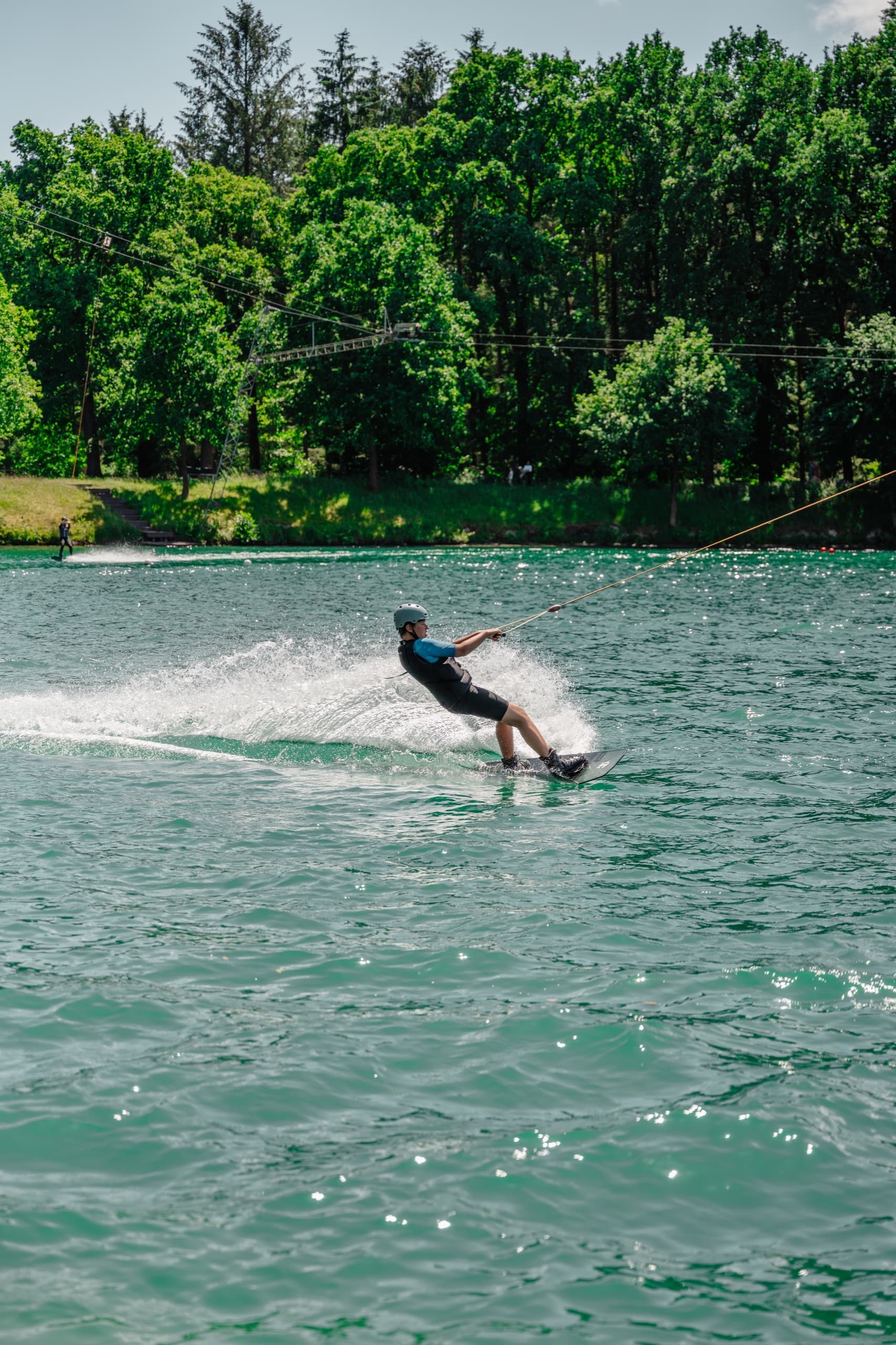 Wakeboarderin in schwarzem Wetsuit surft über den türkis-grünen See beim Schloss Dankern Wakepark