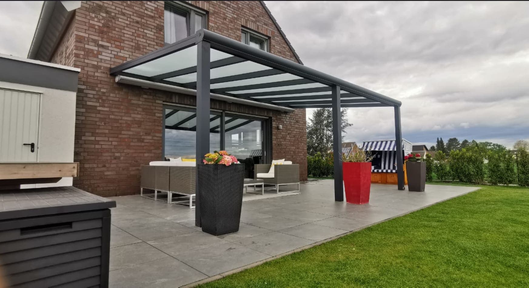 Fertige anthrazitgraue Terrassenüberdachung mit Glasdach über Lounge-Bereich und Strandkorb am Backsteinhaus