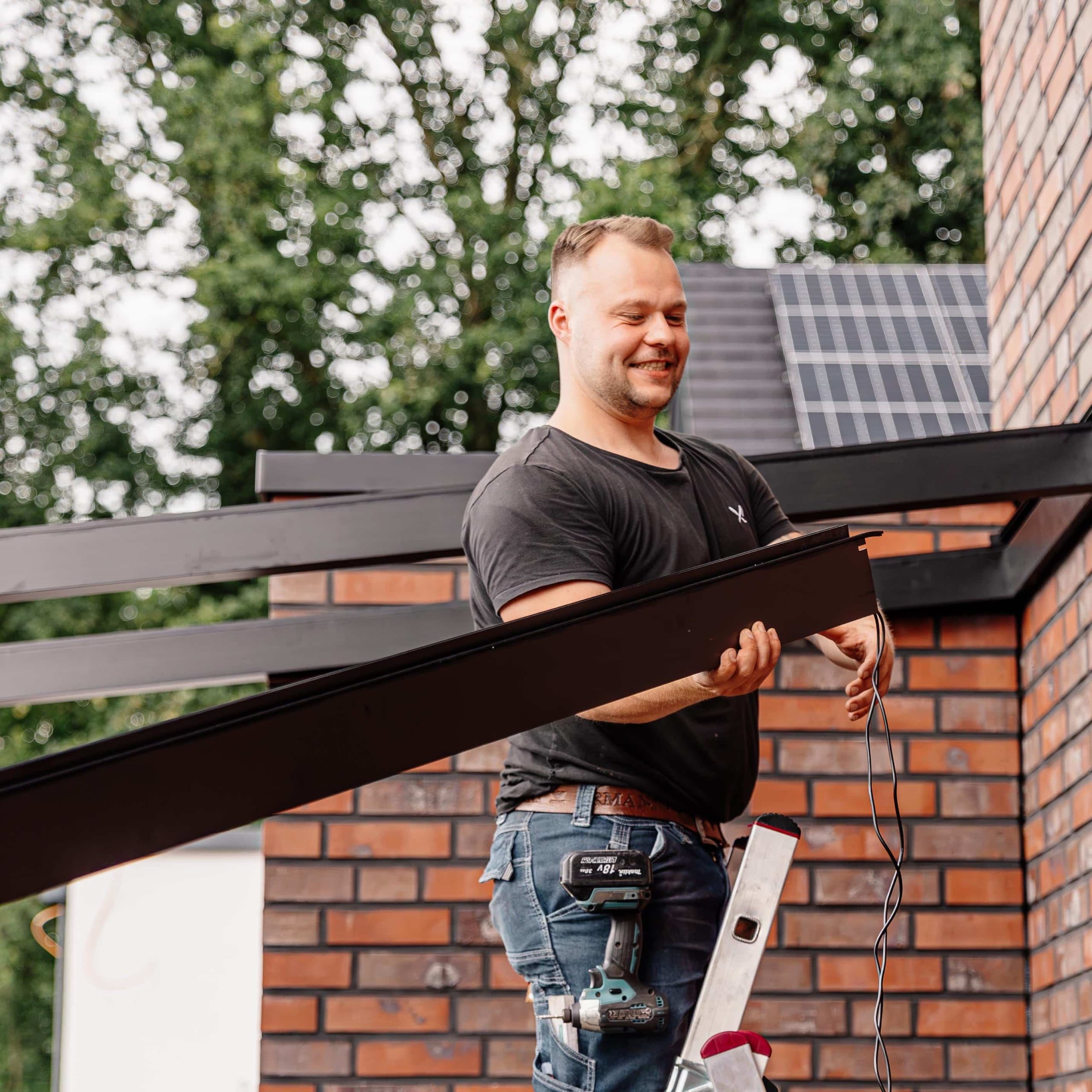 Terravista Meppen Monteur lächelt bei Montage von anthrazitfarbenem Aluminium-Überdachungsrahmen auf Leiter